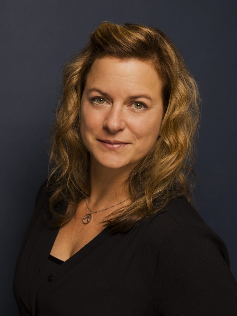 Deborah Thummes Porträt einer Frau mit lockigem, blondem Haar in einem dunklen Hemd vor neutralem Hintergrund.